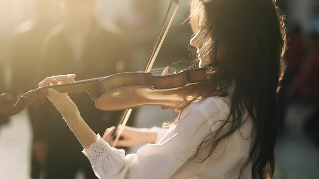 Young street musician playing classical wooden violin on the urban city street. Talented violinist with incredible sunset on the background.