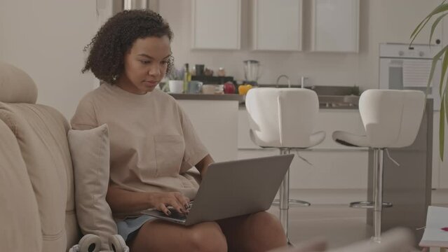 Medium slowmo of Biracial girl in early 20s sitting on sofa in living room studying on laptop