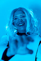 Futuristic concept, a young woman with illuminated virtual reality glasses on a blue background reaching out her hand