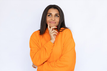 Portrait of dreamy Latin American woman touching face. Thoughtful young model with long hair in orange hoodie looking away, smiling and imagining good things. Dream concept.