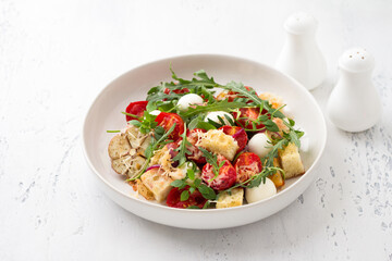 Baked panzanella, traditional Italian salad with tomatoes, mozzarella, parmesan, crispy croutons, arugula and basil on a light blue background