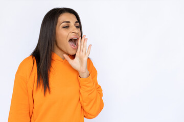 Portrait of screaming Latin American woman. Happy young model with long hair in orange hoodie looking away, shouting with hand near mouth. Studio shot, announcement concept.