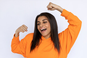 Obraz na płótnie Canvas Portrait of happy Latin American woman dancing. Smiling young model with long hair in orange hoodie looking away, raising hands and singing. Happiness, entertainment concept.