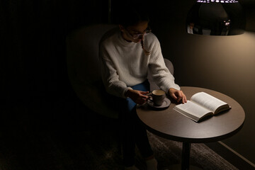 Woman reading a book in her room at night with a coffee. Concept of reading novels at night.