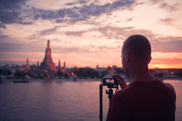Man photographing cityscape at sunset. Tourist with camera on tripod against Wat Arun temple in Bangkok, Thailand....