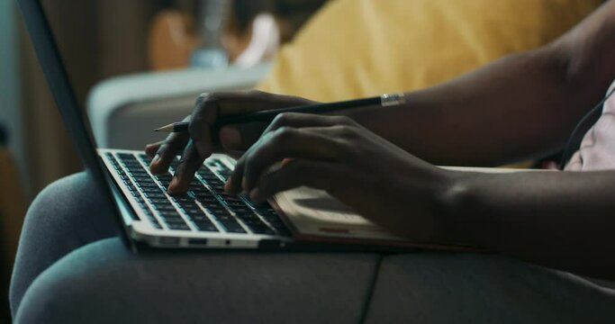 Close-up of hands of African-looking boy holding pencil and typing on laptop keyboard. Man with beard and curly hair is having video conference with colleagues. He is wearing headband and headphones.