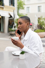 Stylish african american woman talking on cellphone and holding cup of coffee in outdoor cafe in Treviso.