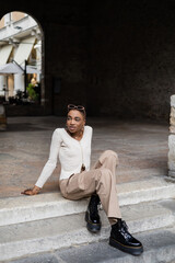 Stylish african american woman with sunglasses sitting on stairs on urban street in Treviso.