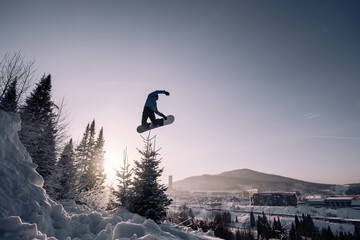 Snowboarder making high big air jump with grab in clear blue sunny sky during sunset above fir...