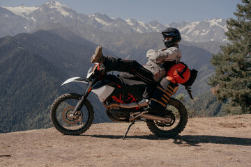 Motorcyclist man wearing helmet and moto clothes sitting in pose of rest on motorcycle against the backdrop of beautiful mountain range
