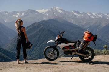 Young woman biker wearing dreadlocks holding helmet standing on vacation in moto trip near motorbike with bags on long road against stunning mountain scenery