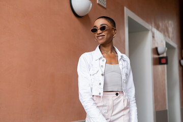 Smiling african american woman in sunglasses and white jacket standing on urban street.