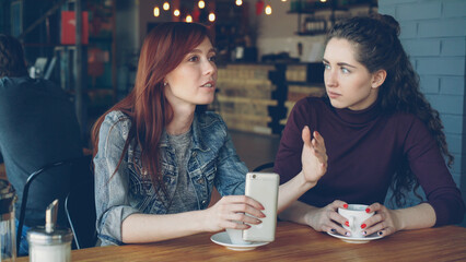 Two attractive women in casual clothes are having girl time in cafe talking and holding coffee cup and smartphone. Nice cafe interior, customers and modern furniture are visible.