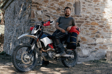 Fototapeta na wymiar motorcyclist sitting on motorcycle with dry bag and wardrobe trunks, resting in moto trip near stone medieval building in the mountains