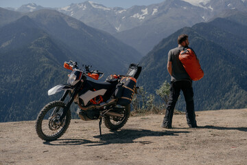 motorcyclist on motorcycle trip arriving at rest stop holding dry bag on his shoulder admiring a...