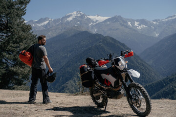 Fototapeta na wymiar Biker on motorcycle trip arriving at rest stop holding dry bag on his shoulder admiring a stunning mountain view