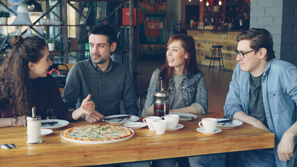 Cheerful young friends are talking and gesturing sharing news while sitting at table in modern cafe together. Big pizza, cups and plates, tables and chairs are visible.