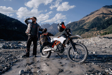 motorcyclist standing near dirt motorcycle in long mountain journey on a mountain river, holding hand covering his face from the bright sun on a summer day