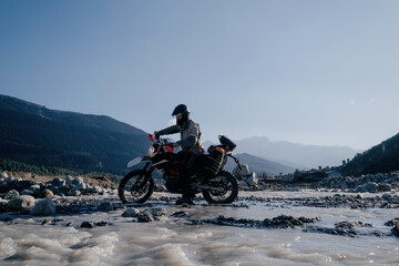 motorcyclist wearing a helmet rides an off-road motorcycle along ford of mountain river in beautiful landscape