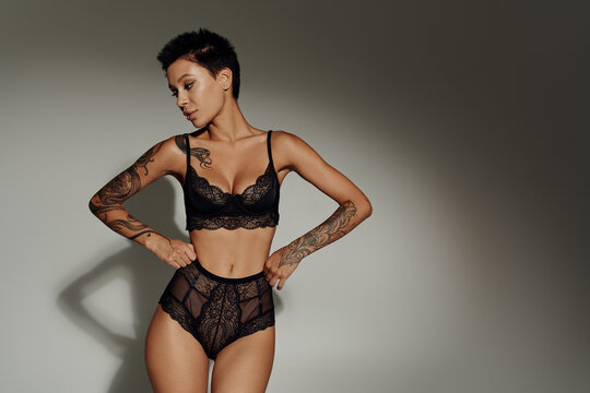 young tattooed woman in black lace lingerie standing with hands on hips on grey background with shadow.