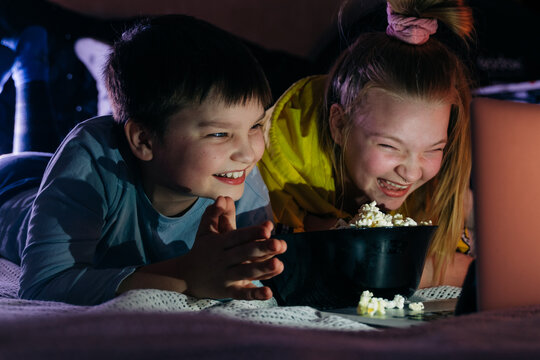 Overjoyed Kids sister brother laughing spend weekend free time together at home on couch bed eating popcorn. teen children watching video cartoon use remote control on laptop have fun. Movie night.5