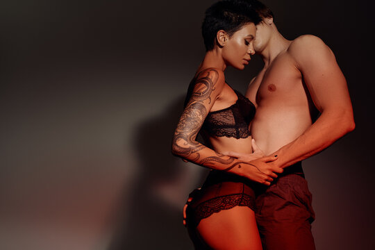 side view of seductive tattooed woman in lingerie near shirtless man on grey background.