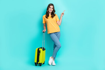 Full length photo of sweet shiny girl dressed yellow t-shirt walking holding luggage pointing empty space isolated teal color background