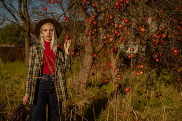 Fashionable confident woman wearing trendy autumn outfit with hat, green checkered blazer, red turtleneck, jeans, holding leather backpack, posing outdoor, near apple tree. Copy, empty space for text
