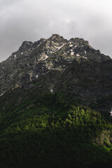 juicy green valley with rivers in grass and trees at the foot of snow-capped rocky mountains with waterfalls, voluminous fluffy thunderclouds through which sunlight breaks through