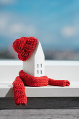 Miniature house in a red scarf and hat on on the windowsill. The concept of passive house heating. Thermal insulation of a building or dwelling. Energy crisis.