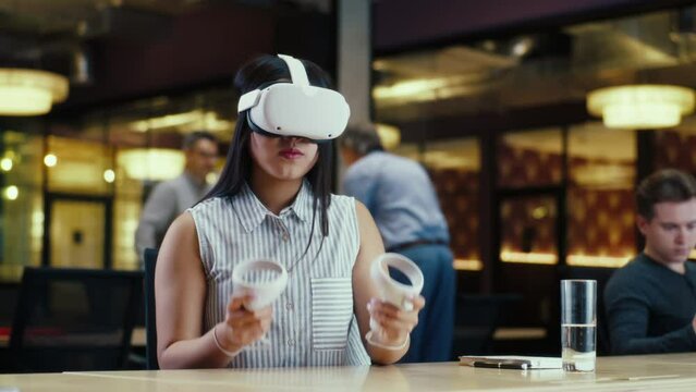 Asian woman wearing VR headset and using wireless controllers, gesturing, watching data in virtual reality. Work in modern office of hi-tech company with diverse colleagues. Cyberspace technology