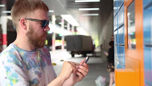 Man with smartphone enters parcel code on screen and picks up parcel from automated postal machine. Man holds smartphone using automatic self-service mail terminal closeup.