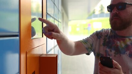 Man with smartphone enters parcel code on screen and picks up parcel from automated postal machine. Man holds smartphone using automatic self-service mail terminal closeup.