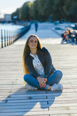 A long haired girl posing in a city outdoors summer,autumn evening.Girl sitting on the wooden floor.Front view.Blurred background green city park trees.Klaipeda,Lithuania.09-02-2022.