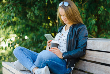Obraz premium A smiling modern young woman sitting on bench in green park on summer day and talking on smartphone with coffee cup.Girl sitting with a phone and coffe at park.Klaipeda,Lithuania.09-02-2022.