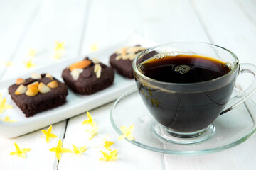 Hot americano black coffee in clear cup on white plate with chocolate brownie cake beside place on old white table and beautiful flower around