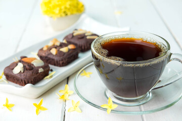 Hot americano black coffee in clear cup on white plate with chocolate brownie cake beside place on old white table and beautiful flower around
