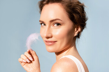 Smiling real  woman holding white feather near glowing perfect face skin. Cosmetology, dermatology....