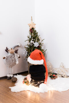 A baby boy is sitting in front of a Christmas tree, he is holding new year lights and wearing Santa Clause red hat