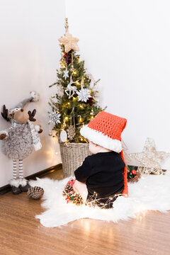 A baby boy is sitting in front of a Christmas tree, he is holding new year lights and wearing Santa Clause red hat