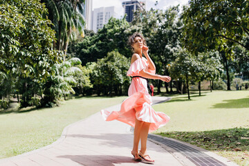 Well-dressed caucasian girl dancing in park in summer morning. Positive young woman in pink dress chilling on nature background.