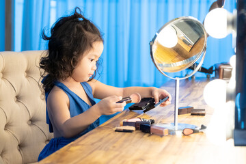 Cute asian toddler girl in blue dress trying makeup blush in front of the mirror.