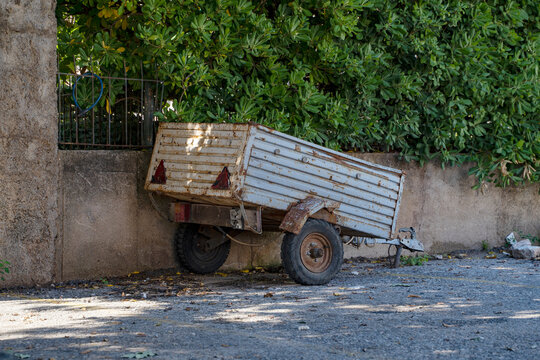 A metal cargo trailer stands in the yard of a house against a background of concrete and green foliage. Cargo cart . Trailer for transporting heavy loads.