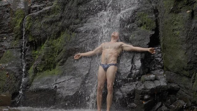 A sexy male athlete, with a naked torso, long black hair, stands under a waterfall, in a mountain river. slow motion. 