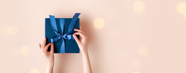 Female hands with festive minimalistic manicure tie a bow on blue gift box. Surprise preparation. Christmas Eve. Xmas and New Year postcard. Black Friday sales, Birthday celebration party concept