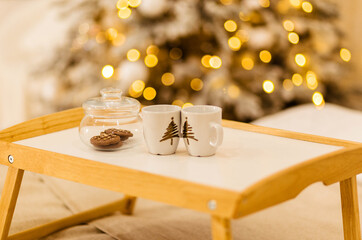 A wooden tray stands on the bed against the backdrop of Christmas lights. On the tray is a jar of cookies and two cups.