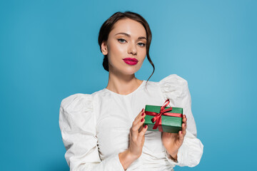 brunette young woman in white blouse holding wrapped present and pooing at camera isolated on blue.