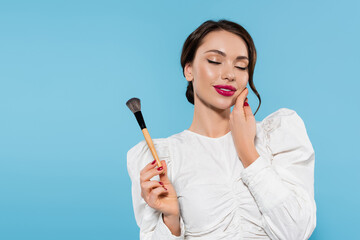 joyful young woman in white blouse holding cosmetic brush and touching cheek isolated on blue.