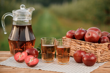 Two glasses with apple juice and basket with apples on wooden table with natural orchard background. Vegetarian fruit composition