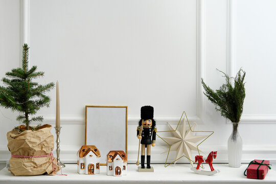 Christmas decor. Nutcracker, candles in the form of houses, a rocking horse and a natural Christmas tree stand on a white shelf. An empty frame for text stands on a shelf